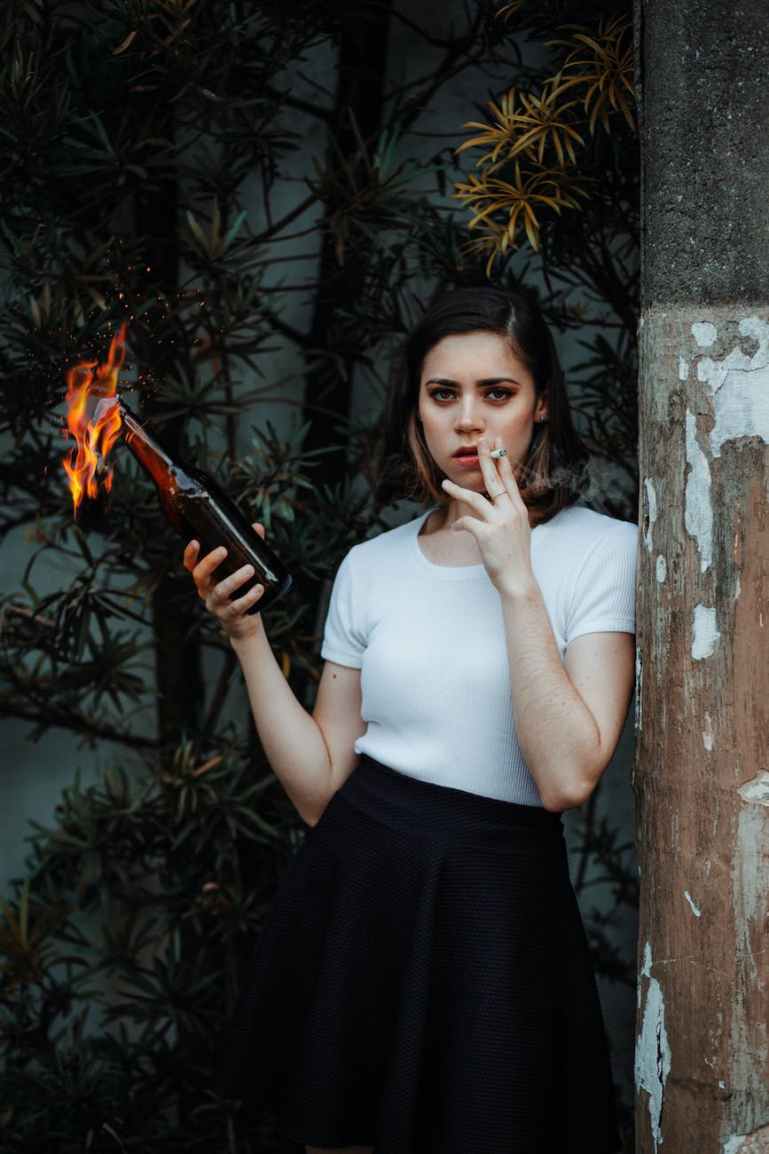 photo of woman leaning on pole while holding molotov cocktail and smoking a cigarette