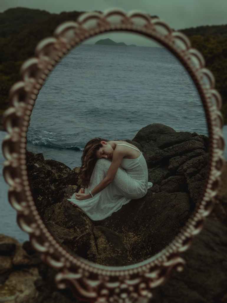 reflection of woman in white dress sitting on brown rock near body of water